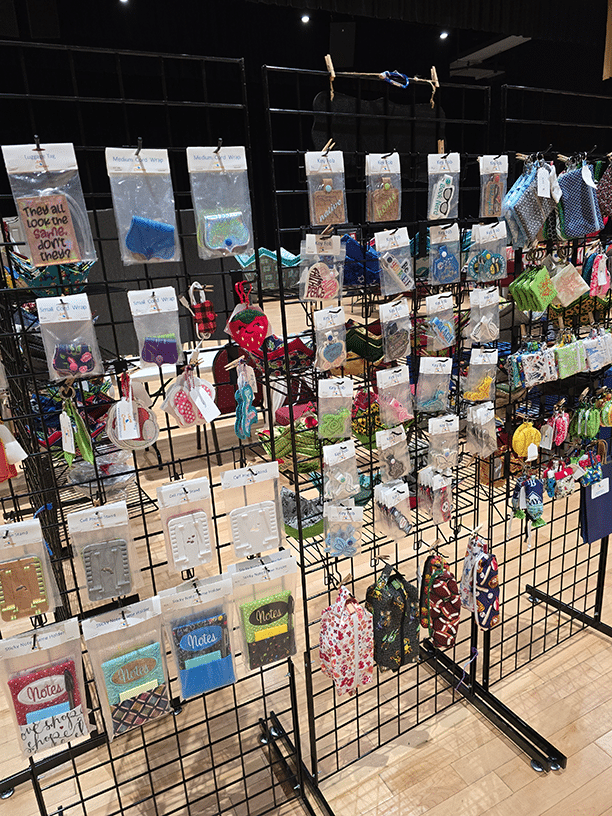 Craft fair display of handmade keychains, notebooks, patches, and kitchen accessories on wire grid panels.