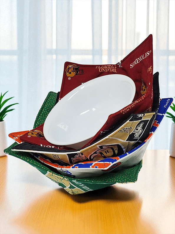 Stack of handmade microwave bowl cozies with college team fabrics holding a white bowl on a kitchen table.